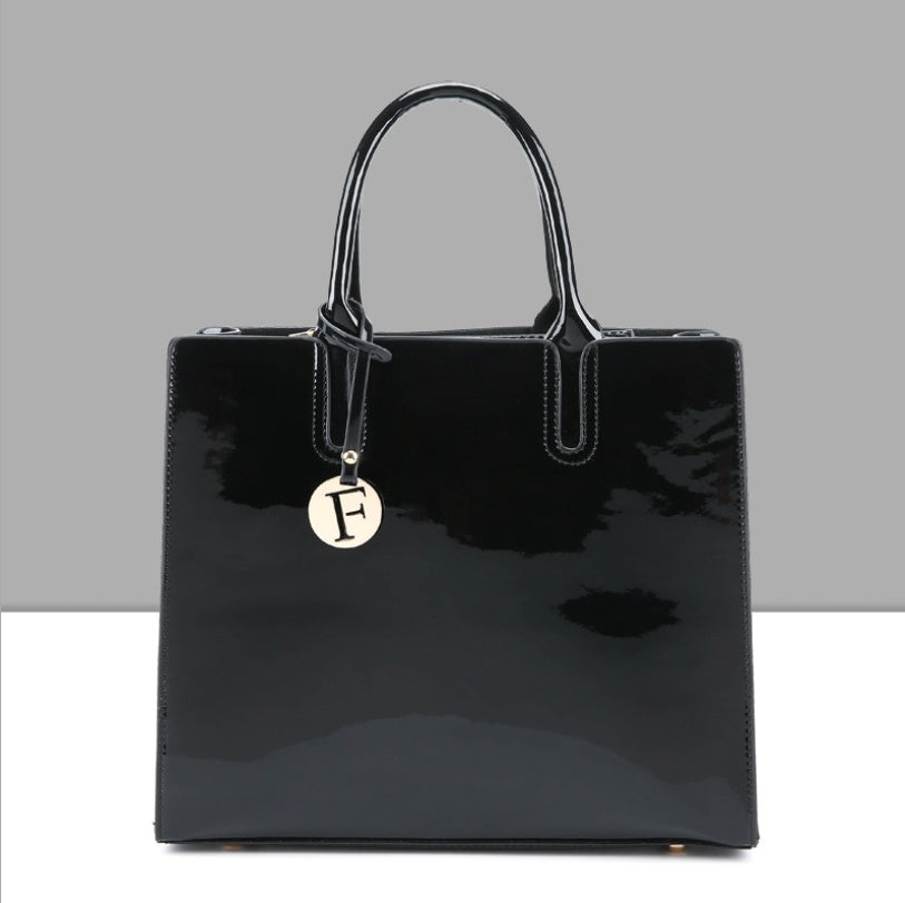 Black patent leather handbag with a gold charm on a gray background